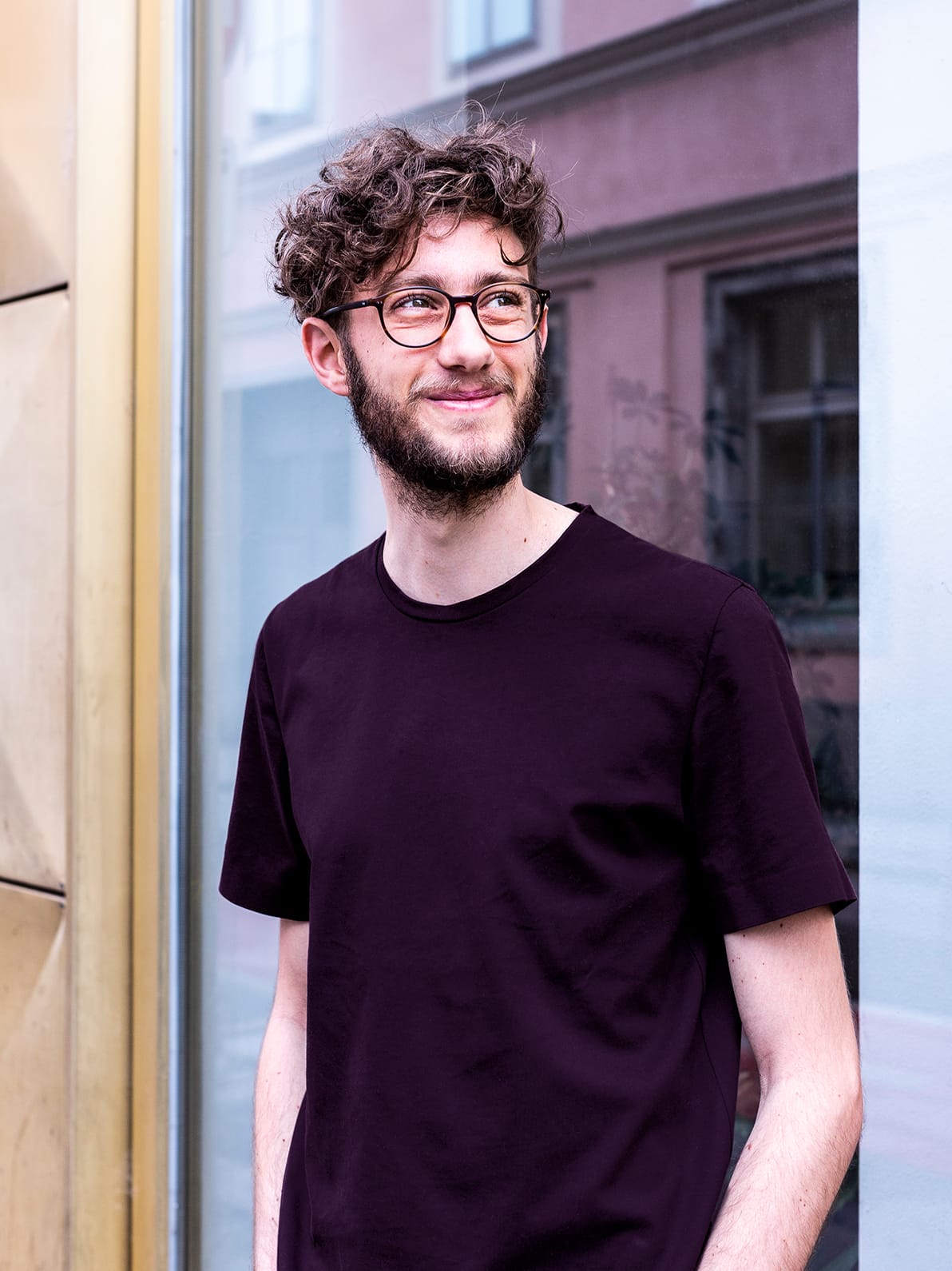 Ein junger Mann mit lockigem Haar, Bart und Brille, trägt ein weinrotes T-Shirt und lächelt nach rechts.