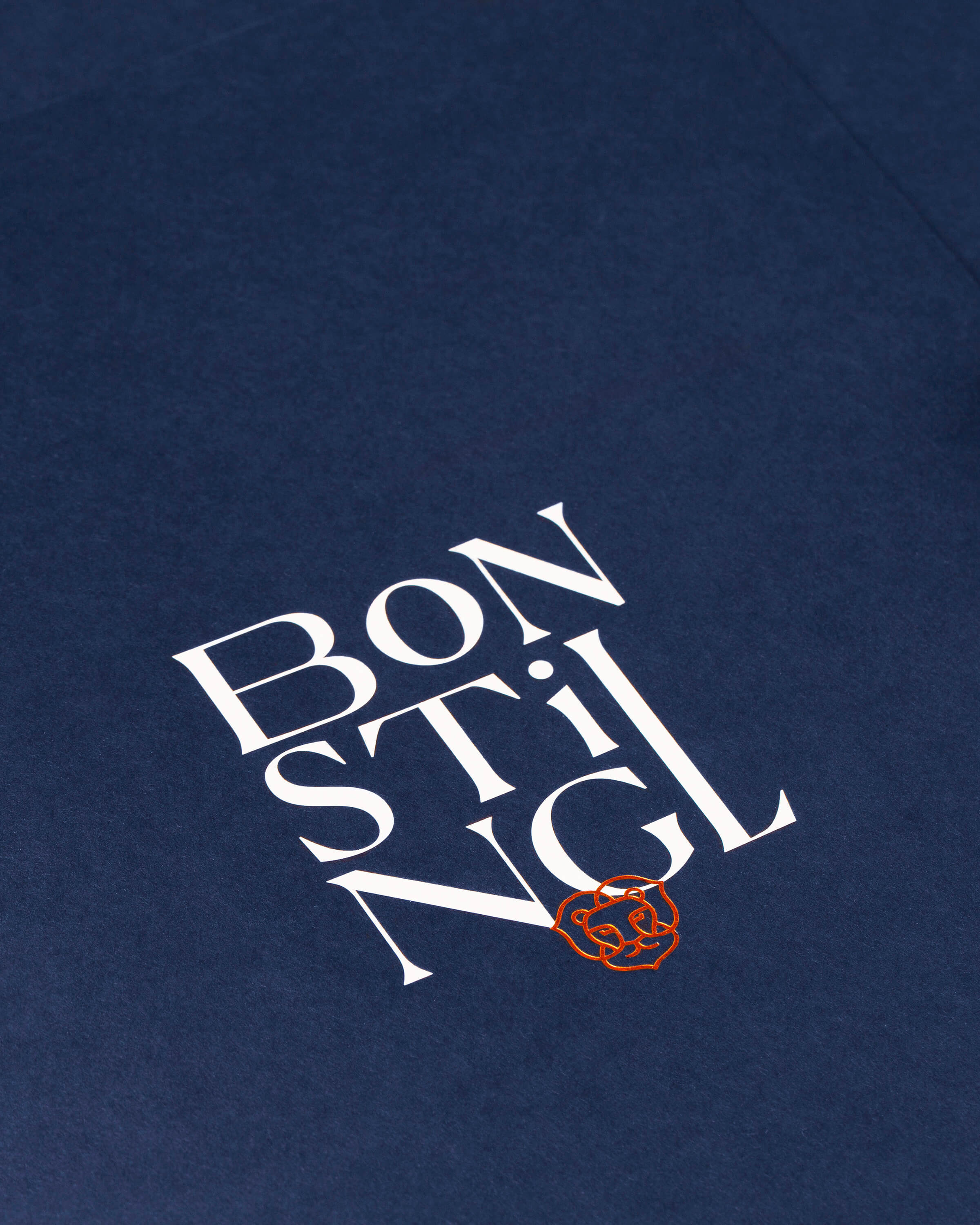 Nahaufnahme des Logos "BON STIL NGL" in Weiß mit einem orangefarbenen Bärengesicht auf einem dunkelblauen Stoff.